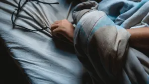 Eine Hand greift nach einer Brille in einem ungemachten Bett im Halbdunkel.